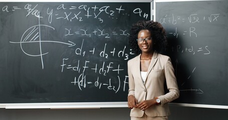 portrait of young african american female teacher in glasses standing at board, writing with chalk m