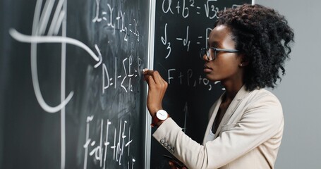 African American young woman teacher at school writing formulas and mathematics laws on blackboard. School concept. Female math lecturer in glasses explaining physics laws. Education.