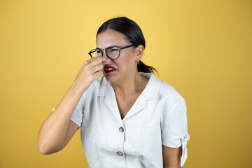 Wall Mural - Beautiful woman over yellow background smelling something stinky and disgusting, intolerable smell, holding breath with fingers on nose.