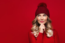 Pretty Blonde Woman With Curly Hair Touching Her Chin, Wears Red Wool Turtleneck And Hat, Isolated On Red Studio Wall, Enjoys The Winter Weather, Thinking And Looking At Blank Space For Advertising