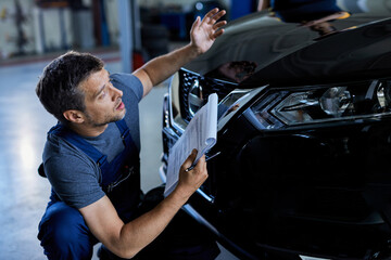 Car mechanic reading data from clipboard while examining vehicle hood in auto repair shop.