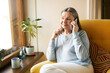 © shurkin_son - Age, leisure, technology and electronic gadgets. Cheerful casually dressed senior woman with gray hair relaxing indoors sitting in armchair, taking to friend on mobile, drinking coffee by window