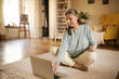 © shurkin_son - Technology, gadgets and aging concept. Barefoot European female pensioner using portable computer at home, siting on carpet, surfing internet or watching online yoga video tutorial for aged people
