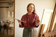 © shurkin_son - Mature people, aging, hobby and leisure. Indoor image of good looking concentrated senior woman in apron using palette to mix oil pigments, applying paint on canvas using brush, making painting