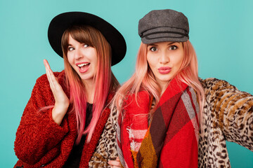 Close up studio portrait of two pretty cute models with pink hairs in stylish leopard print faux fur coat and trendy hat on turquoise. Surprise face. Winter fashionable outfit.