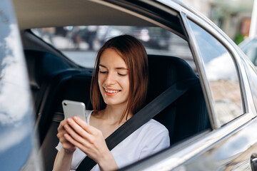 Beautiful, young woman uses a smartphone and smiles while sitting in the back seat of a car