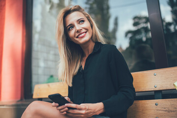 Caucasian cheerful hipster girl with perfect blonde hair taking rest on bench in city enjoying free time for cellphone networking, happy female with cute candid smile holding mobile technology