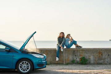 Wall Mural - Outdoor shot of couple of travelers sitting near the broken car with open hood and smoking engine, man and woman looking tired, frustrated while waiting for assistance