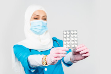 Wall Mural - friendly Muslim doctor or nurse in a hijab, mask, gloves offering a pill to the patient on a gray background