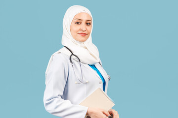 Wall Mural - friendly, Muslim doctor or nurse woman in a hijab with a stethoscope writes a prescription on pills on a blue background.