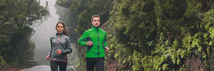 Runners jogging outside on wet trail running in the rain in autumn fall outdoor cold season. Panoramic of couple athletes training together.