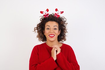 Wall Mural - Positive Young arab woman with curly hair wearing christmas headband standing on white  smiles happily, glad to receive pleasant news from interlocutor, keeps hands together. People emotions concept.