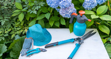 Gardening Tools In Blue - Pruning Shears, Spray Bottle, Scissors, And A Hat With Gloves Against A Background Of Blue Flowers As Preparation For Gardening Work
