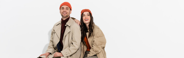 Poster - panoramic shot of stylish woman in trench coat looking up near trendy man in beanie hat on white