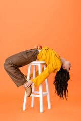 Wall Mural - trendy woman in autumn outfit with apples on belly lying on white stool and touching boots isolated on orange