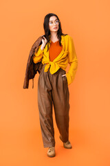 Wall Mural - full length of young woman in autumn outfit holding leather jacket and standing with hand in pocket on orange