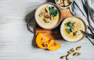 two plates of pumpkin soup next to greens pumpkin and bread croutons on a white wooden table top view close up