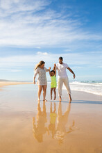 Happy Family Couple And Little Girl Enjoying Walking And Activities On Beach, Kid Holding Parents Hands, Jumping And Hanging. Front View. Family Outdoor Activities Concept