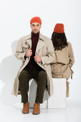 Sticker - stylish man in beanie hat touching trench coat near woman sitting on white