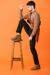 Poster - side view of stylish man in autumn outfit and glasses standing with leg on wooden stool on orange