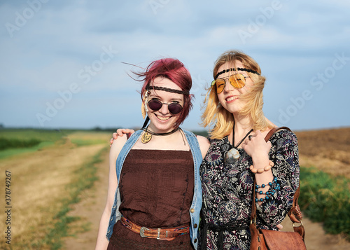 Fotomural Two hippie women, wearing boho style clothes, walking on dirt road on green field, having fun