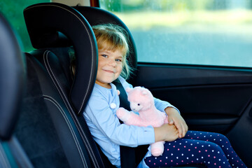 Adorable toddler girl sitting in car seat, holding plush soft toy and looking out of the window on nature and traffic. Little kid traveling by car. Child safety on the road. Family trip and vacations