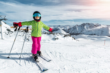 Active adorable preschooler caucasian kid girl portrait with ski in helmet, goggles and bright suit enjoy winter extreme sport activities. Little child skiing on luxury alpine resort in mountains