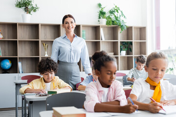 selective focus of teacher looking at camera near multiethnic pupils during lesson in school