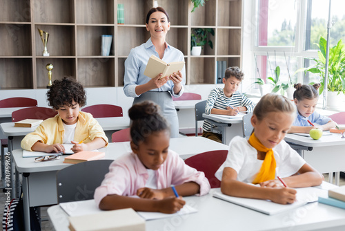 Fotografía teacher holding book and looking at camera while multicultural pupils writing di