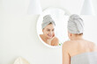 © amixstudio - Portrait of  young girl with  towel on head in white bathroom looks and touches her face in the mirror and enjoys youth and hydration. Natural beauty, home care for problem skin