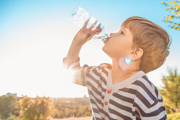 A beautiful child sitting on the grass drinks water from a bottle in the summer at sunset. Boy quenches his thirst on a hot day