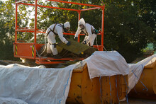 Professional Asbestos Removal. Two Men In Protective Suits Are Removing Asbestos Cement Corrugated Roofing, Bringing It From The Roof Into A Container