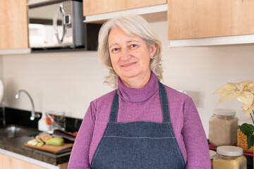 Lovely Senior grandmother with cooking apron smiling in camera at kitchen table, inside. Togetherness, multi-generation family, support concept.