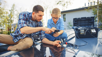 Father and Son Installing Solar Panels to a Metal Basis. They Work with Wiring on a House Roof on a Sunny Day. Boy is Looking at a Manual on a Smartphone. Father Connects the Plugs.