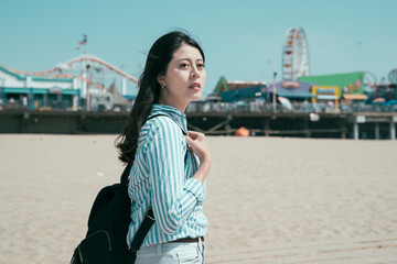 Canvas Print - beautiful asian japanese woman backpacker walking on sandy beach outdoor on sunny day. side view of young relax charming female tourist with bag enjoy ocean with blurred amusement park in background.