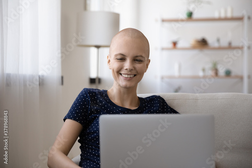 Happy young hairless sick woman struggling with oncology relax on sofa at home using modern laptop. Smiling bald ill female suffering from cancer look at computer screen, watching video online.