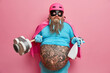 © wayhome.studio  - Funny man superhero foolishes around and does cleaning holds detergent and plunger has thick beard with clothing pins big tattooed belly isolated on pink studio background. Housekeeping concept