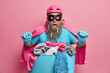 Fat bearded superhero man poses over basin full of dirty laundry wears pink cloak and helmet stares at camera with surprised face expression isolated on pink background. Housekeeping concept