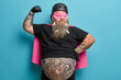 © WHstudio Leushin N - Funny bearded adult man with thick beard pretends being powerful superhero raises arm and shows muscles demonstrates power ready to protect you wears pink mask cloak poses against blue background