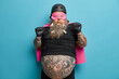 © WHstudio Leushin N - Scared fearful man has big tattooed belly wears superhero suit for costume party stares shocked at camera poses with raised hands against blue background. Frightened male wears cape and helmet