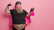 © WHstudio Leushin N - Horizontal shot of powerful superhero shows muscles and holds detergent or household supplies ready to clean everything, works in cleaning service poses against pink background with blank space.