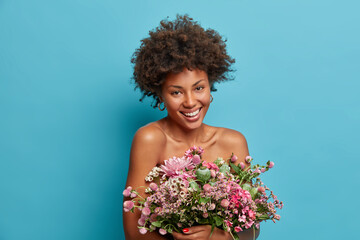 Indoor shot of glad smiling woman has healthy skin and natural curly hair poses bare shoulders with present bouquet of flowers against blue background enjoys pleasant aroma. Holiday concept.