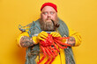 © WHstudio Leushin N - Stupefied bearded sailor holds big octopus and smokes pipe poses with fishing net around neck has tattooed arms isolated on yellow background. Boatswain carries big sea creature stands indoor