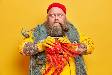 Stupefied bearded sailor holds big octopus and smokes pipe poses with fishing net around neck has tattooed arms isolated on yellow background. Boatswain carries big sea creature stands indoor