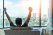 Business achievement concept with happy businessman relaxing in home office or hotel room, resting and raising fists with ambition looking forward to city building urban scene