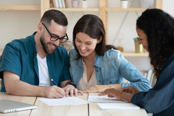 Happy young married couple signing contract, making successful deal, smiling young husband wearing glasses putting signature on legal documents, purchasing new house, taking loan or mortgage