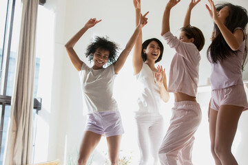 Energetic young mixed race diverse female friends in pajamas having fun, dancing to favorite music, jumping on bed, entertaining on sleepover girls party, enjoying weekend fun time together.