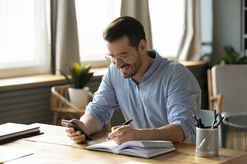 Wall Mural - Smiling young Caucasian man sit at desk at home office workplace look at cellphone screen watch webinar making notes. Happy millennial male use smartphone summarize plan writing in journal.