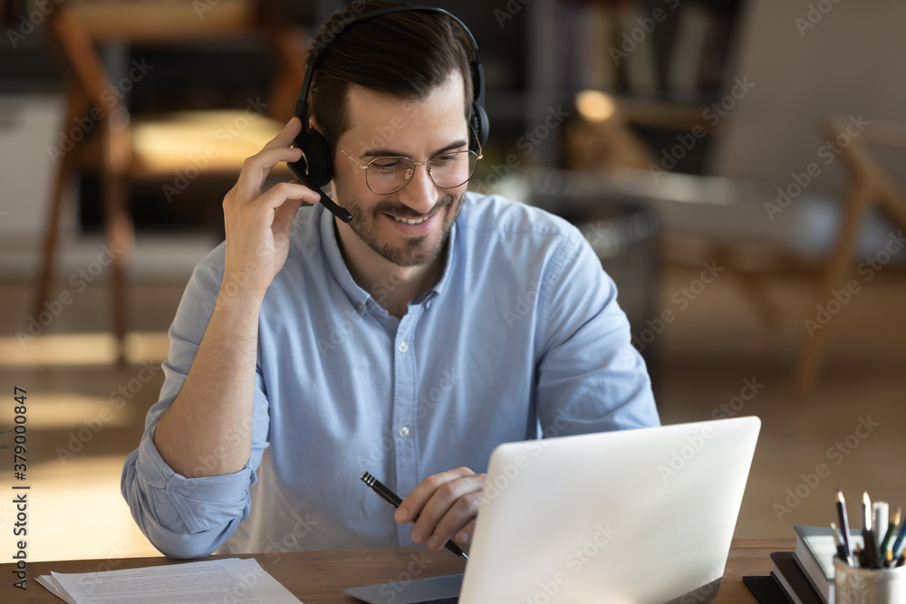 Smiling millennial Caucasian man in headset look at laptop screen work online from home office. Happy young man in headphones watch webinar take course or training on web. Distant education concept. - obrazy, fototapety, plakaty 
