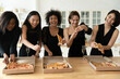 Happy young festive mixed race pretty women best friends in classic black dresses having fun at party, eating delivery italian fast food pizza, drinking champagne, enjoying meeting together indoors.
